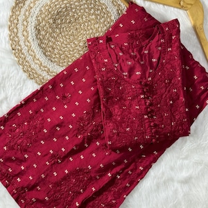 May include: A maroon embroidered kurta and palazzo set with gold detailing. The set is folded and laid out on a white fluffy surface.