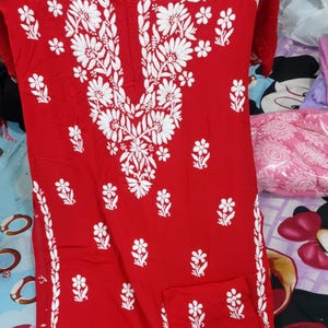 Puede incluir: Una t&uacute;nica roja brillante de manga larga con intrincados bordados florales blancos alrededor del cuello y a los lados. La prenda tiene un escote en V y una silueta recta, con tela roja a juego doblada en la esquina inferior derecha.