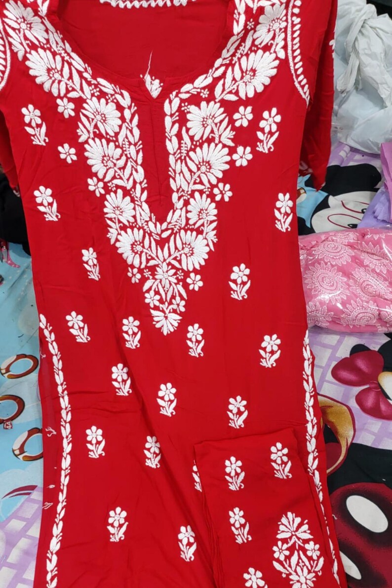 Puede incluir: Una t&uacute;nica roja brillante de manga larga con intrincados bordados florales blancos alrededor del cuello y a los lados. La prenda tiene un escote en V y una silueta recta, con tela roja a juego doblada en la esquina inferior derecha.