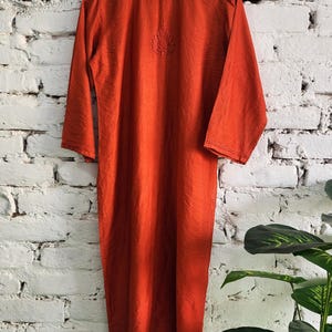 May include: A long, burnt orange dress with long sleeves hangs on a wooden hanger against a white brick wall. The dress features a subtle embroidered design near the neckline. The fabric appears to have a slight sheen.