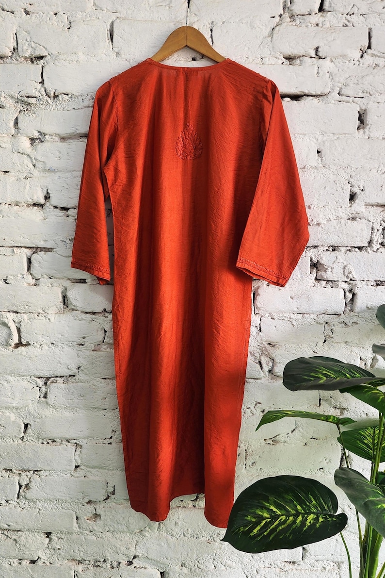 May include: A long, burnt orange dress with long sleeves hangs on a wooden hanger against a white brick wall. The dress features a subtle embroidered design near the neckline. The fabric appears to have a slight sheen.
