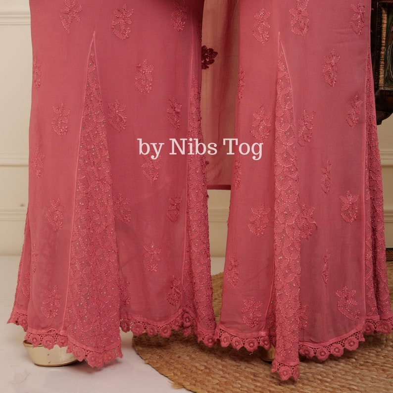 May include: A pair of pink palazzo pants with embroidered floral details and a lace trim. The pants have a wide leg and a flowing silhouette. The text "by Nibs Tog" is visible on the pants.