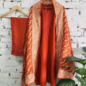 Puede incluir: Un conjunto salwar kameez bordado naranja, que incluye una túnica, pantalones y un chal. La túnica y los pantalones son de color naranja liso, mientras que el chal presenta intrincados diseños color crema. El conjunto cuelga de una percha de madera contra una pared de ladrillos blancos.