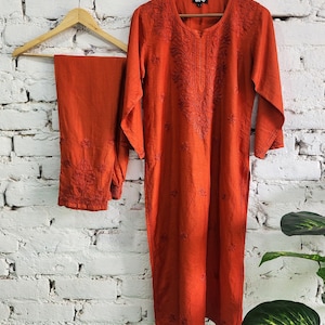 May include: A burnt orange embroidered kurta and matching pants set hanging on a wooden hanger against a white brick wall. The kurta features detailed floral embroidery around the neckline and sleeves. The pants also have embroidered details.