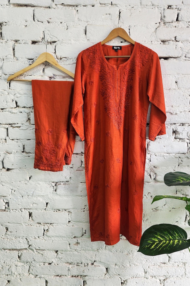 May include: A burnt orange embroidered kurta and matching pants set hanging on a wooden hanger against a white brick wall. The kurta features detailed floral embroidery around the neckline and sleeves. The pants also have embroidered details.