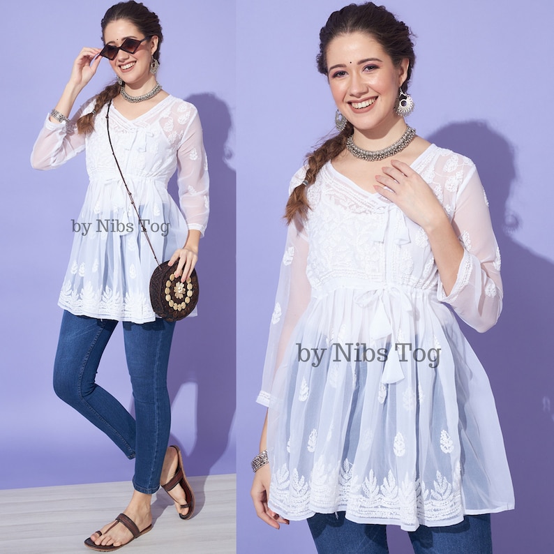 May include: White cotton tunic top with embroidery and a tie at the waist. The top is worn with blue jeans and brown sandals. The text "by Nibs Tog" is visible on the top.