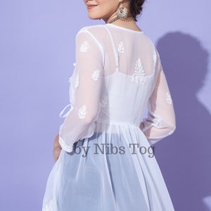 May include: White sheer tunic top with embroidered floral details and a tie at the waist. The top is made of a lightweight fabric and has a flowy silhouette. The text "by Nibs Tog" is visible on the bottom of the top.
