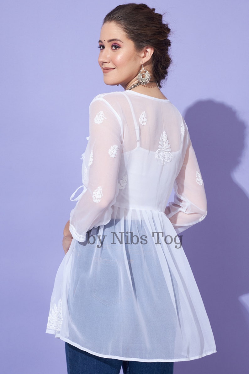 May include: White sheer tunic top with embroidered floral details and a tie at the waist. The top is made of a lightweight fabric and has a flowy silhouette. The text "by Nibs Tog" is visible on the bottom of the top.