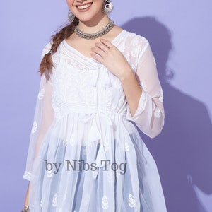 May include: White sheer tunic top with embroidered floral details and a tie at the waist. The top is made of a lightweight fabric and has a flowy silhouette. The text "by Nibs Tog" is printed on the front of the top.