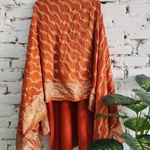 May include: Burnt orange shawl with intricate gold-colored embroidery. The fabric drapes over a matching garment, creating a layered effect. The design features a repeating wave pattern and floral details, against a white brick wall.