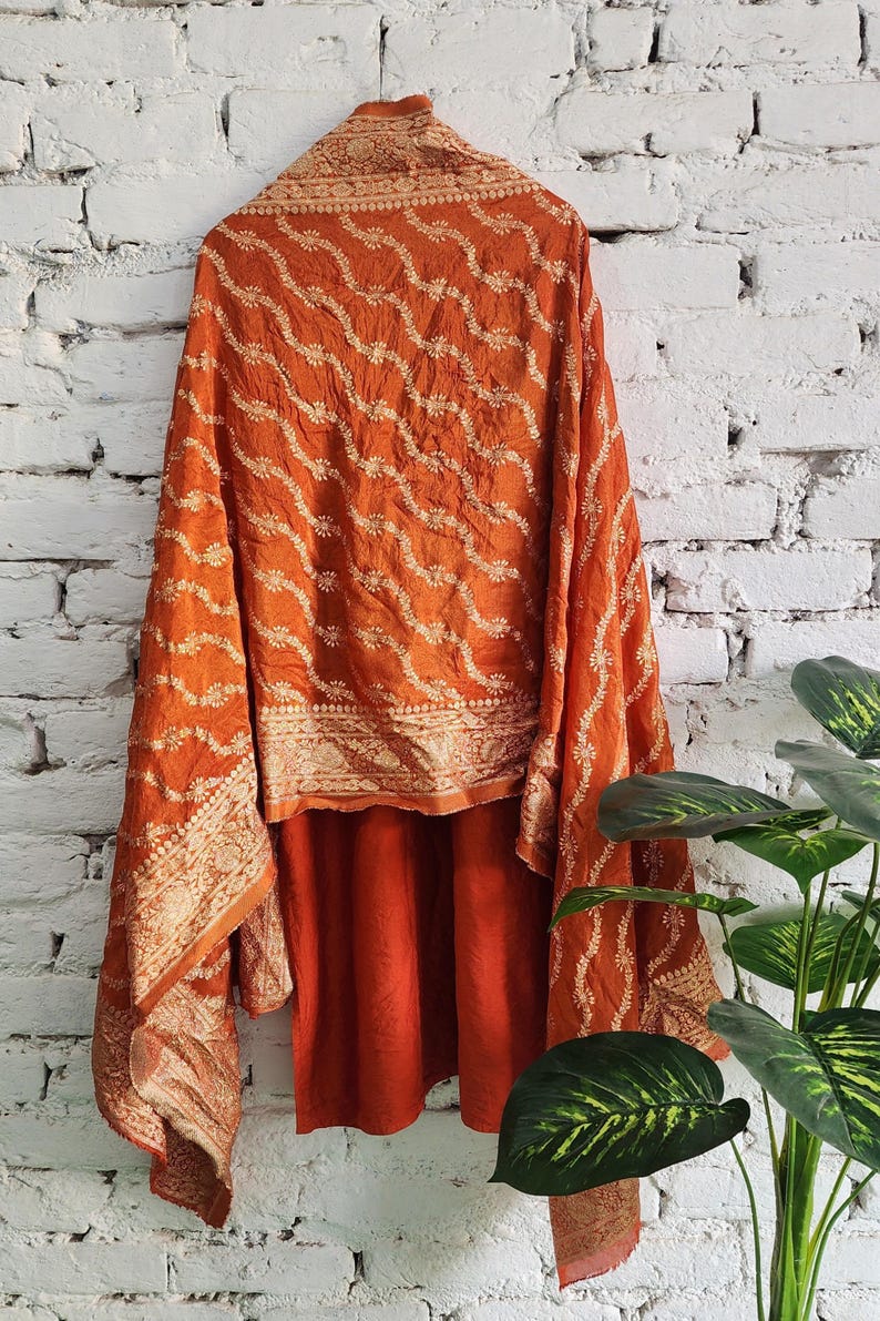 May include: Burnt orange shawl with intricate gold-colored embroidery. The fabric drapes over a matching garment, creating a layered effect. The design features a repeating wave pattern and floral details, against a white brick wall.