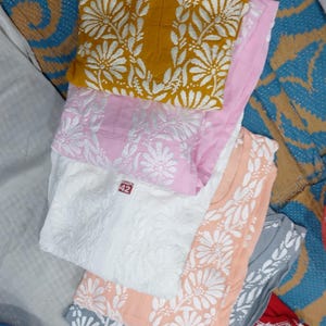 Puede incluir: Una colecci&oacute;n de seis kurtas plegadas en amarillo mostaza, rosa claro, blanco, melocot&oacute;n, gris y rojo. Cada prenda presenta un patr&oacute;n de bordado floral blanco en la parte delantera y una peque&ntilde;a etiqueta roja cerca del cuello con el n&uacute;mero 42.
