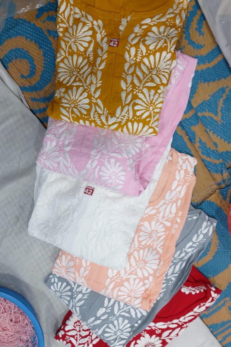 Puede incluir: Una colecci&oacute;n de seis kurtas plegadas en amarillo mostaza, rosa claro, blanco, melocot&oacute;n, gris y rojo. Cada prenda presenta un patr&oacute;n de bordado floral blanco en la parte delantera y una peque&ntilde;a etiqueta roja cerca del cuello con el n&uacute;mero 42.