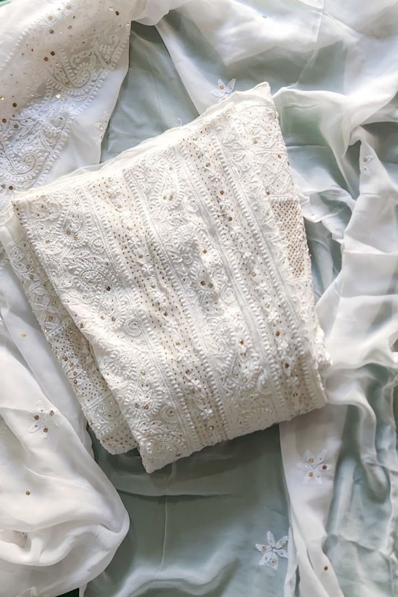 May include: White embroidered fabric with gold sequins. The fabric is folded in half and laid on a white and green surface.
