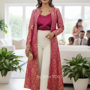 Puede incluir: Una mujer con un abrigo largo rosa y dorado estampado, top magenta y pantalones blancos. Sostiene un clutch dorado y lleva joyas doradas. El texto "by Nibs TOG" es visible.