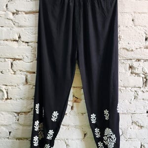 Puede incluir: Pantalones negros con cintura el&aacute;stica y silueta de pierna c&oacute;nica. La parte inferior de las piernas est&aacute; decorada con patrones de bordado floral en blanco, que incluyen peque&ntilde;as flores individuales y un grupo m&aacute;s grande cerca del dobladillo.