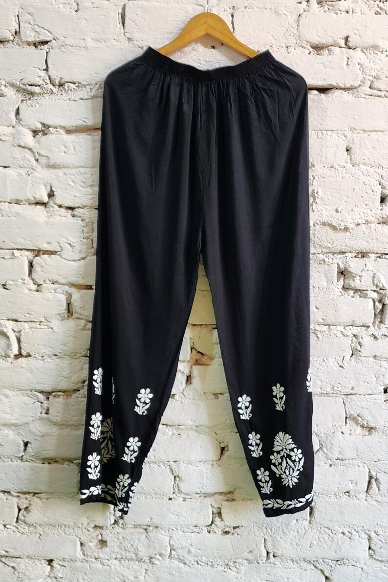 Puede incluir: Pantalones negros con cintura el&aacute;stica y silueta de pierna c&oacute;nica. La parte inferior de las piernas est&aacute; decorada con patrones de bordado floral en blanco, que incluyen peque&ntilde;as flores individuales y un grupo m&aacute;s grande cerca del dobladillo.