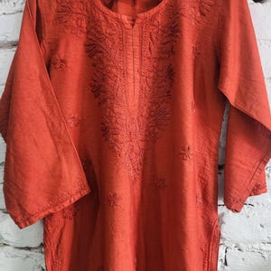 May include: An orange tunic with three-quarter sleeves and intricate floral embroidery around the neckline and down the front. The tunic is hanging on a wooden hanger against a white brick wall. The brand name "Nidhi Vyas" is visible on the top.