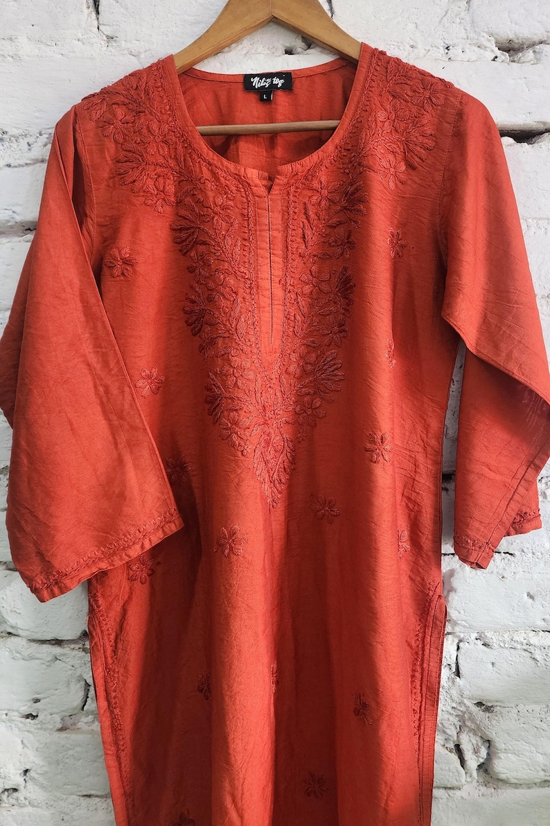 May include: An orange tunic with three-quarter sleeves and intricate floral embroidery around the neckline and down the front. The tunic is hanging on a wooden hanger against a white brick wall. The brand name "Nidhi Vyas" is visible on the top.