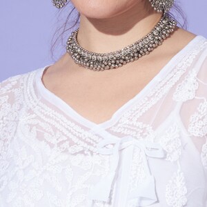 May include: A woman wearing a white, embroidered top with a silver necklace and earrings. The necklace is made of small, round beads and the earrings are large and circular.