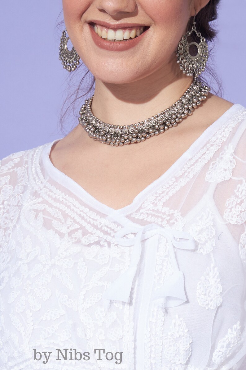 May include: A woman wearing a white, embroidered top with a silver necklace and earrings. The necklace is made of small, round beads and the earrings are large and circular.