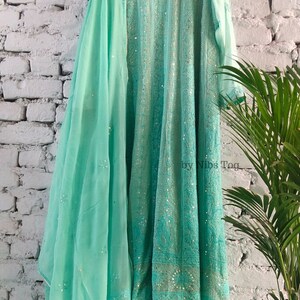 May include: A turquoise blue embroidered dress with a matching dupatta. The dress has a round neckline and three-quarter sleeves. The dupatta is sheer and has a delicate floral pattern. The dress is adorned with sequins and beads. "by Nibs Tog" is printed on the dress.