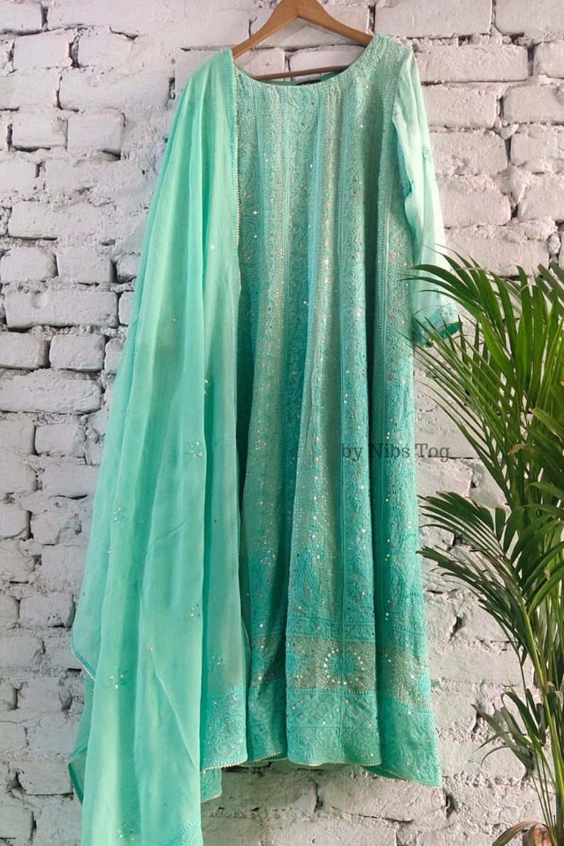 May include: A turquoise blue embroidered dress with a matching dupatta. The dress has a round neckline and three-quarter sleeves. The dupatta is sheer and has a delicate floral pattern. The dress is adorned with sequins and beads. "by Nibs Tog" is printed on the dress.