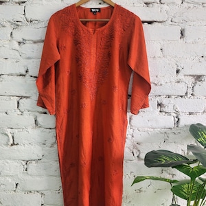 May include: An orange, long-sleeved dress with intricate embroidery around the neckline and down the front. The dress is hanging on a wooden hanger against a white brick wall. The dress has a relaxed fit and a knee-length hemline.