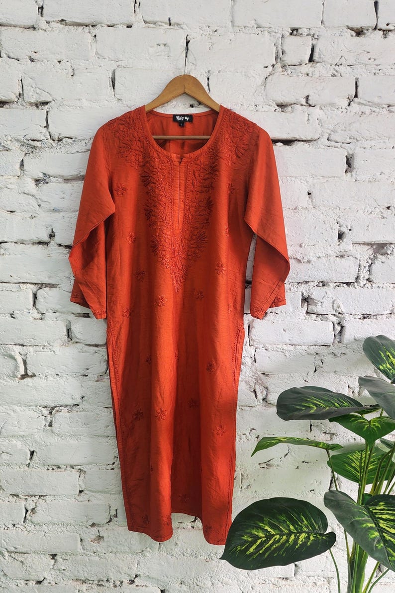 May include: An orange, long-sleeved dress with intricate embroidery around the neckline and down the front. The dress is hanging on a wooden hanger against a white brick wall. The dress has a relaxed fit and a knee-length hemline.
