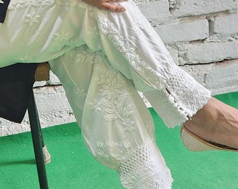 May include: White cotton trousers featuring intricate floral embroidery and a wide, textured lace cuff at the hem. The pants have a tapered silhouette with a gathered design. The text "by Nibs Tog" is visible on the fabric near the knee area.
