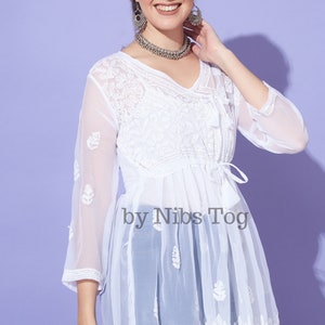 May include: White sheer tunic top with embroidered floral details and a tie at the waist. The top is made of a lightweight fabric and has a flowing silhouette. The text "by Nibs Tog" is printed on the front of the top.