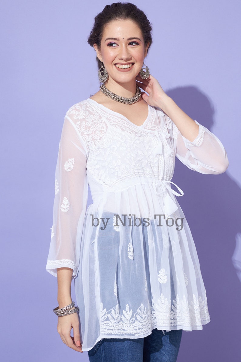 May include: White sheer tunic top with embroidered floral details and a tie at the waist. The top is made of a lightweight fabric and has a flowing silhouette. The text "by Nibs Tog" is printed on the front of the top.
