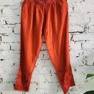 May include: Burnt orange pants hanging on a wooden hanger against a white brick wall. The pants have a loose fit with an elastic waistband and embroidered floral details near the ankles. The fabric has a slight sheen.