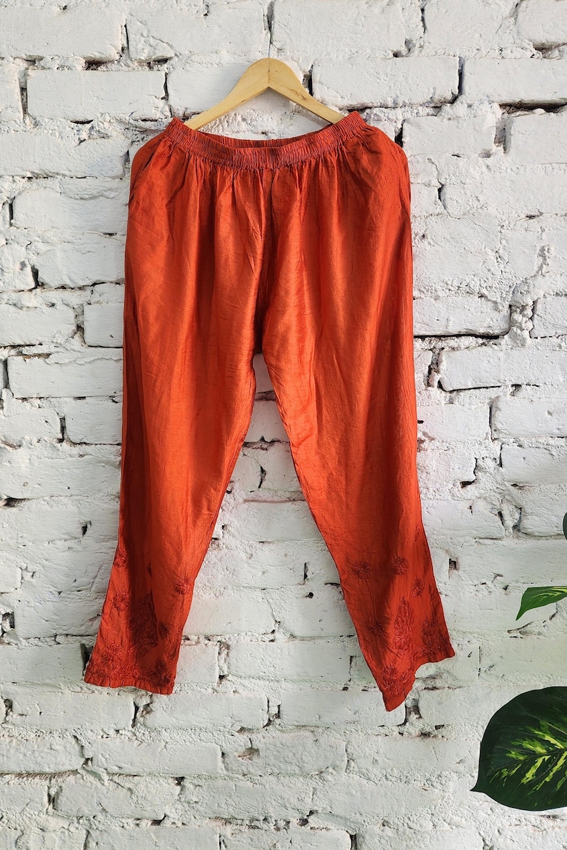 May include: Burnt orange pants hanging on a wooden hanger against a white brick wall. The pants have a loose fit with an elastic waistband and embroidered floral details near the ankles. The fabric has a slight sheen.