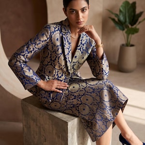 May include: A navy blue and gold brocade blazer and matching midi skirt set featuring a repeating floral and paisley pattern. The blazer has a notched lapel, long sleeves, and a single button closure. The skirt is a straight-cut midi length.