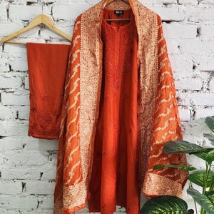 May include: An orange embroidered salwar kameez set, including a tunic, pants, and a shawl. The tunic and pants are a solid orange, while the shawl features intricate white embroidery along the borders. The outfit is displayed on a wooden hanger against a white brick wall.