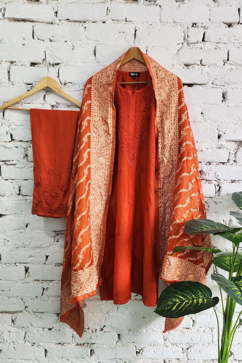 May include: An orange embroidered salwar kameez set, including a tunic, pants, and a shawl. The tunic and pants are a solid orange, while the shawl features intricate white embroidery along the borders. The outfit is displayed on a wooden hanger against a white brick wall.