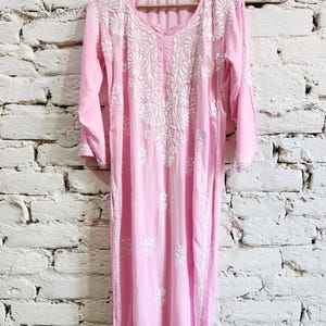 Puede incluir: Una t&uacute;nica de color rosa claro con mangas largas que presenta un intrincado bordado floral blanco alrededor del cuello y en la parte delantera. La prenda tiene cuello redondo, mangas tres cuartos y dobladillo recto, colgada en una percha de madera.