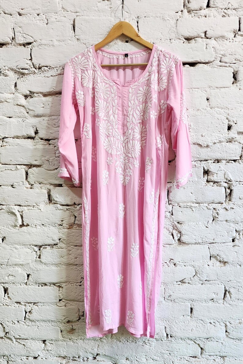 Puede incluir: Una t&uacute;nica de color rosa claro con mangas largas que presenta un intrincado bordado floral blanco alrededor del cuello y en la parte delantera. La prenda tiene cuello redondo, mangas tres cuartos y dobladillo recto, colgada en una percha de madera.