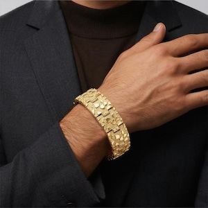 May include: A gold-colored bracelet with a textured, nugget-like design. The bracelet is wide and appears to be made of metal. The bracelet is on a wrist, and the person is wearing a dark suit jacket and a brown turtleneck.