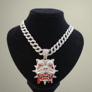 Op de afbeelding: Een zilverkleurige ketting met een grote hanger met een bulldogkop. De hanger heeft rode accenten op de ogen en mond, met een druipend bloedeffect. De ketting is gemaakt van in elkaar grijpende schakels.