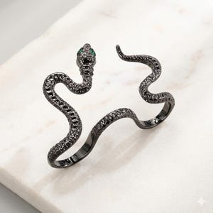 Puede incluir: Un anillo de metal negro con forma de serpiente enroscada y una superficie texturizada similar a escamas. La cabeza de la serpiente presenta dos pequeñas piedras verdes brillantes como ojos. El anillo tiene un diseño abierto y sinuoso.