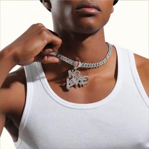 Op de afbeelding: Zilverkleurige ketting met een hanger met de tekst "NO LOVE". De hanger en ketting zijn bezet met kleine, glinsterende steentjes. De ketting wordt gedragen over een witte geribbelde tanktop. De ketting heeft een Cuban link stijl.