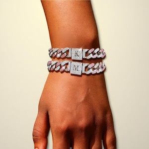 Peut inclure: Deux bracelets de chaîne argentés avec des pierres roses et transparentes. Chaque bracelet a un fermoir carré avec une lettre, l'un avec "K" et l'autre avec "M". Les bracelets sont sur le poignet d'une personne.