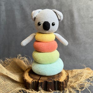 Puede incluir: Un juguete de koala de ganchillo hecho a mano con una pila de anillos coloridos. El koala es gris con una nariz negra y ojos negros pequeños. Los anillos son amarillos, naranjas, verdes y azules, y el juguete se exhibe sobre una base de madera.