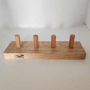 May include: A rectangular light brown wooden display stand featuring four vertical cylindrical pegs arranged in a single row. The front face of the base displays the word Neda in a cursive script, burned into the wood grain surface.