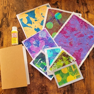 May include: A selection of colourful art prints with abstract designs, a bottle of Mod Podge, and a blank kraft paper notebook. The prints display various colours and patterns, including blue, purple, green, and yellow.