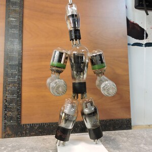 May include: A quirky sculpture made from old vacuum tubes, arranged to look like a person. The tubes are of different sizes and shapes, with black and green details. The figure is set against a wooden backdrop.