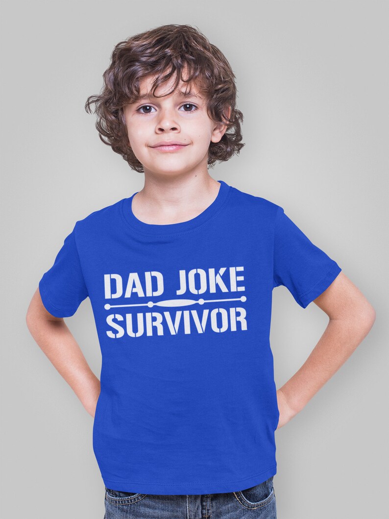 Pu&ograve; includere: Una maglietta blu reale con la scritta bianca "DAD JOKE SURVIVOR". La maglietta &egrave; indossata da un giovane con i capelli castani ricci. La persona sorride e ha le mani sui fianchi.