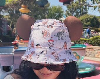 May include: A white bucket hat with a Toy Story print featuring characters like Woody, Buzz Lightyear, and Jessie. The hat has brown ears on the sides.